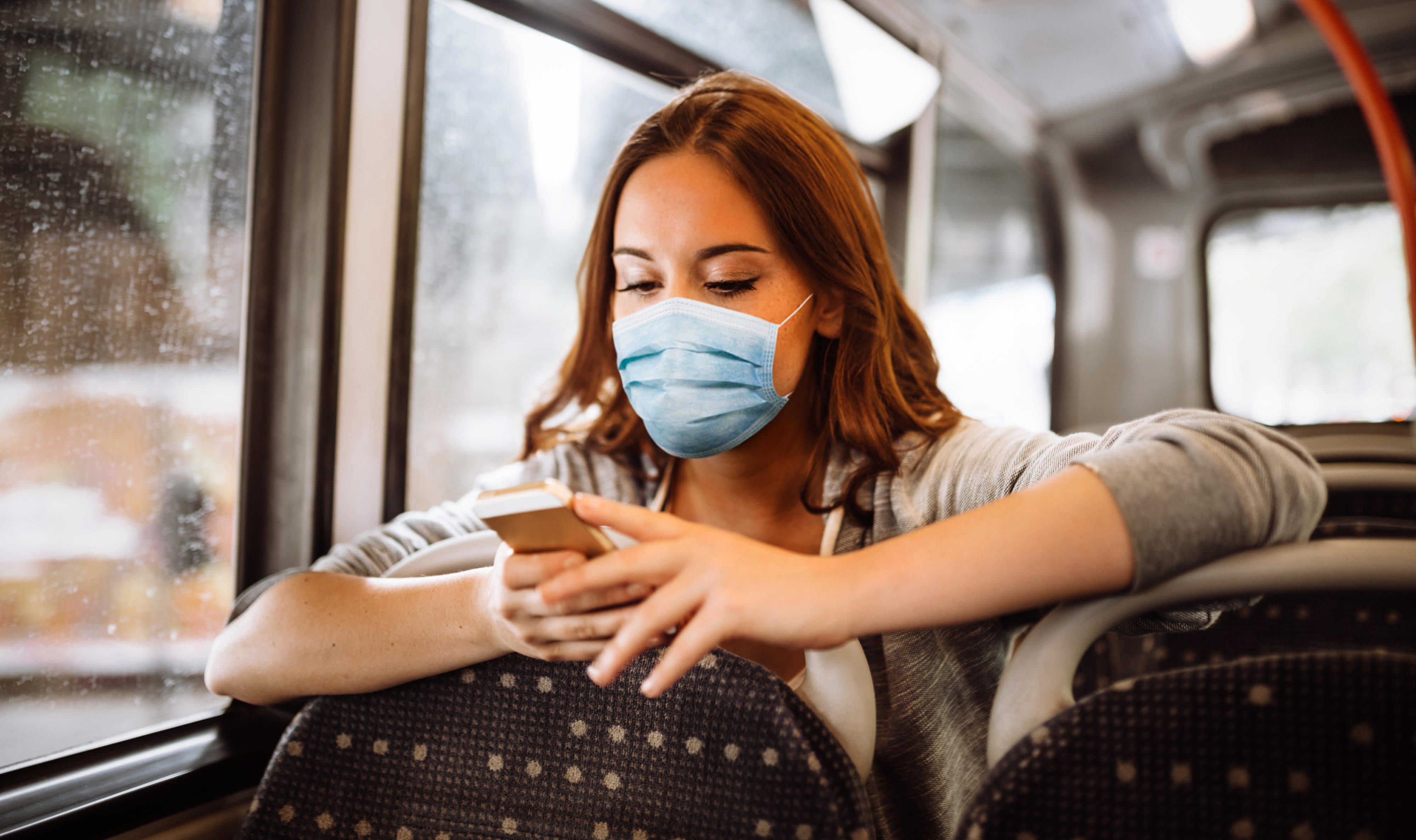Eine junge Frau mit Mundschutz und Smartphone im Bus