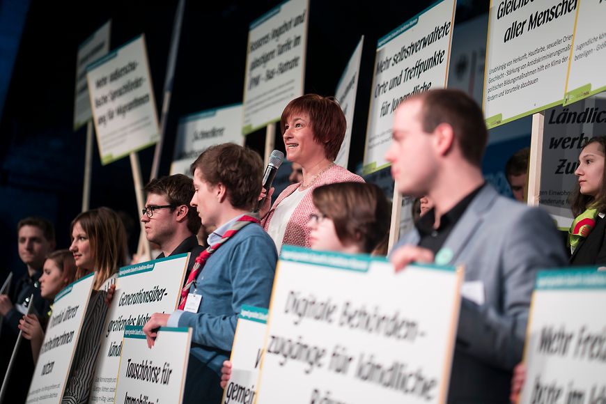 Caren Marks hält gemeinsam mit Jugendlichen Schilder hoch auf der Bühne des Demografiegipfels