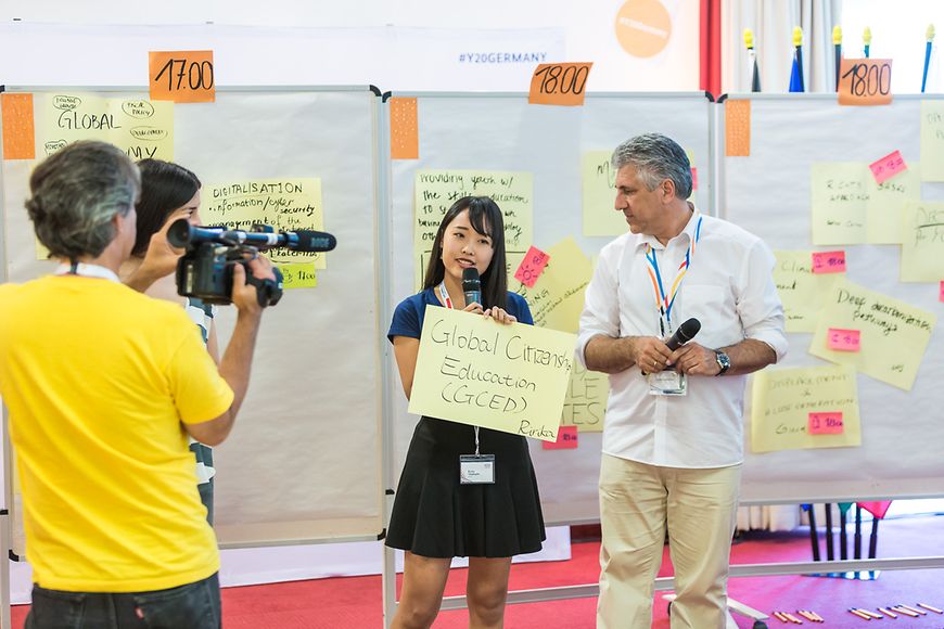 Eine junge Frau präsentiert zum Thema Global Cizizenship Education, ein Kamerateam filmt sie, der Moderator steht daneben