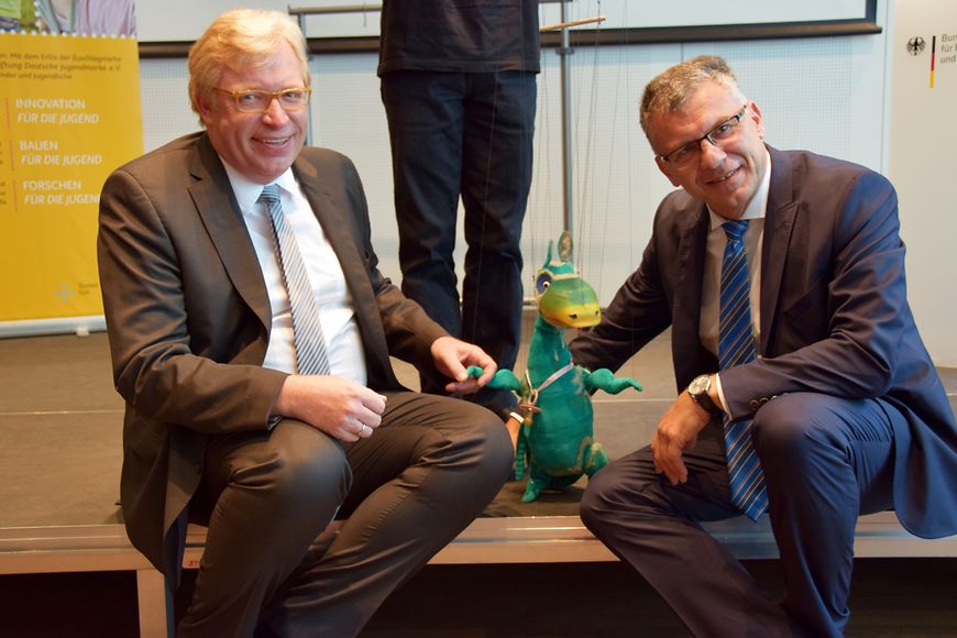 Dr. Ralf Kleindiek und Werner Gatzer sitzen mit der Puppe Urmel in ihrer Mitte auf einer Bühne