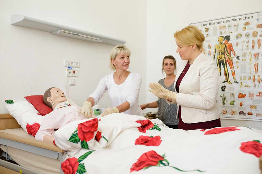 Franziska Giffey steht mit zwei Frauen an einem Bett. Darin liegt eine Puppe zur Übung der Altenpflege.
