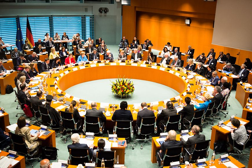 Ein großer, runder Tisch in der Mitte eines Raumes an dem Personen sitzen und Verhandlungen führen