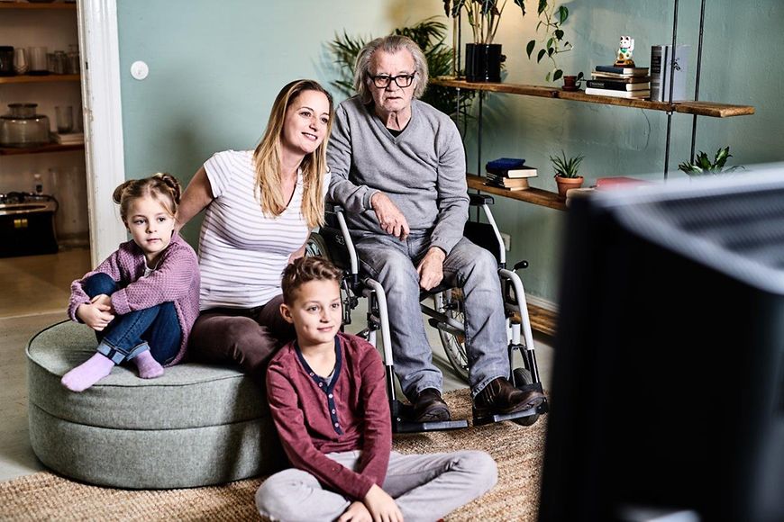 Eine Familie in einem Wohnzimmer vor dem Fernseher, der Großvater sitzt im Rollstuhl