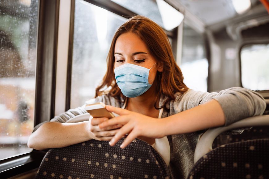 Eine junge Frau mit Mundschutz und Smartphone im Bus