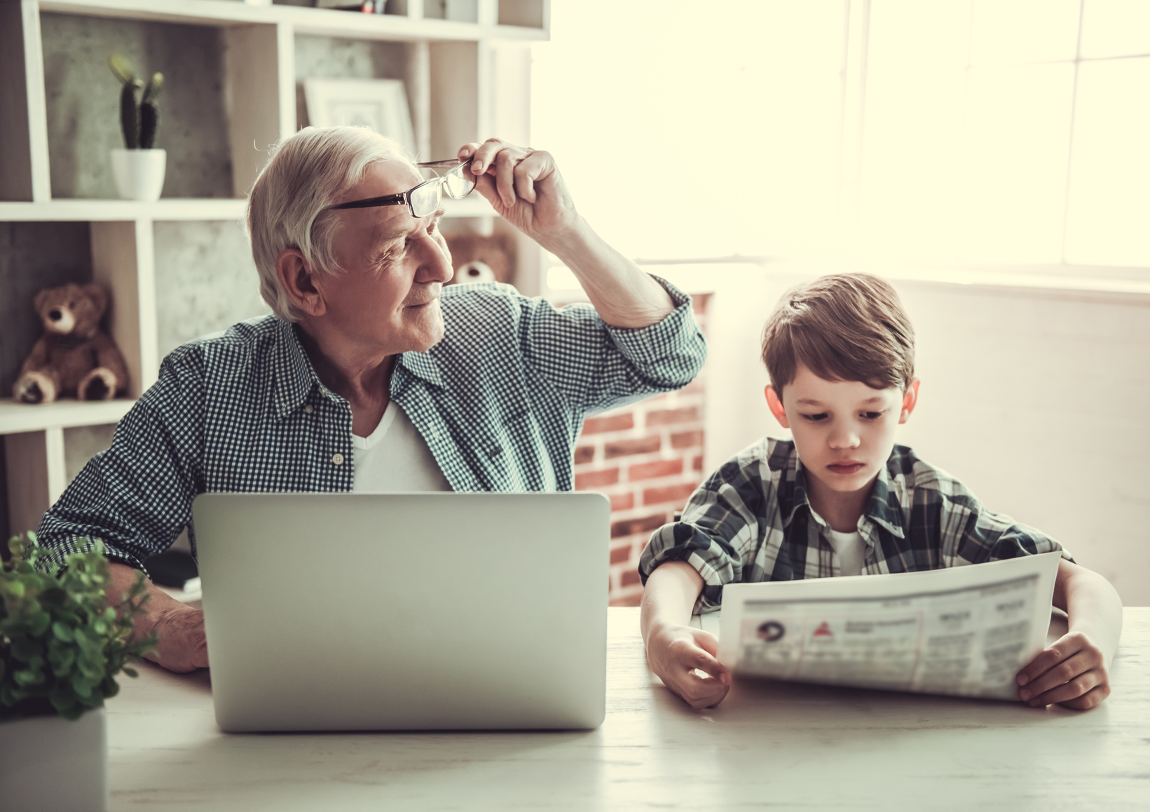 Ein Großvater mit Laptop und sein Enkel mit Zeitung sitzen gemeinsam am Tisch