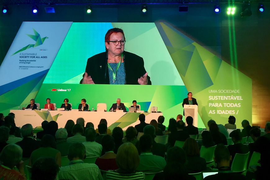 Publikum vor Bühne mit sechs Konferenzteilnehmern, Elke Ferner spricht am Podium, im Hintergrund eine Projektion von Frau Ferner
