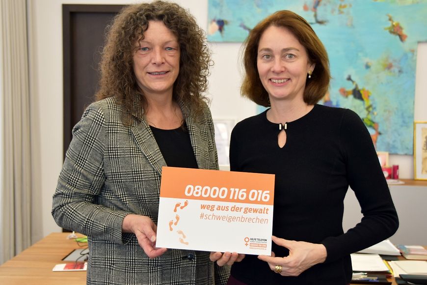 Dr. Katarina Barley und Petra Söchting halten ein Schild mit der Telefonnummer des Hilfetelefons: 08000 116 016