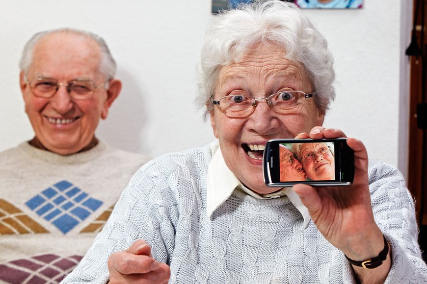 Ein älteres Liebespaar. Die Frau hält ein Smartphone in die Kamera, auf dem ein Foto mit dem küssenden Paar zu erkennen ist.