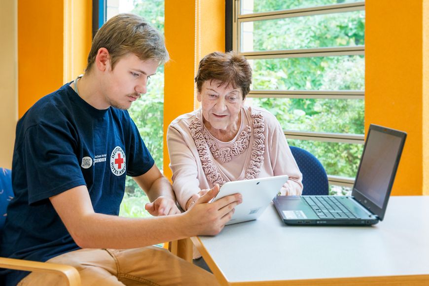 Das Bild zeigt einen Jugendlichen, der einer älteren Dame vor einem Rechner sitzend etwas erklärt