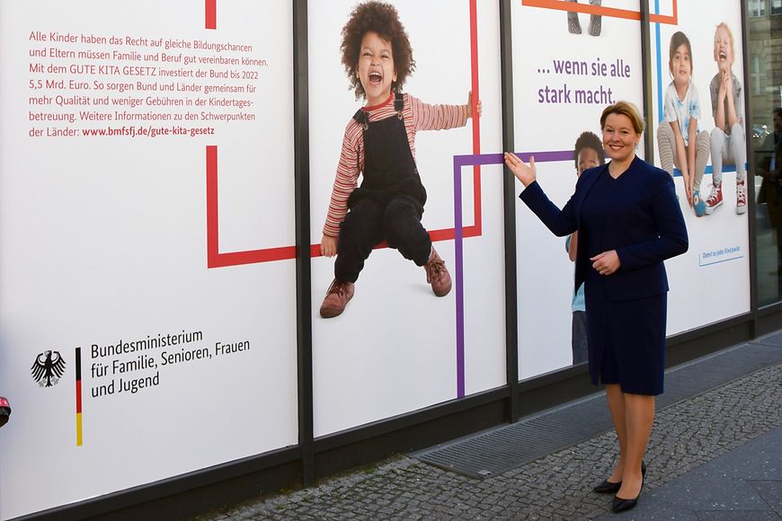 Das Bild zeigt Franziska Giffey vor dem Bundesfamilienministerium, das mit einem neuen Plakat zum Gute-Kita-Gesetz beklebt wurde
