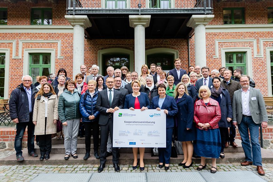 Ein große Menschengruppe steht vor einem Gebäude, versammelt hinter der symbolisch vergrößerten Kooperationsvereinbarung