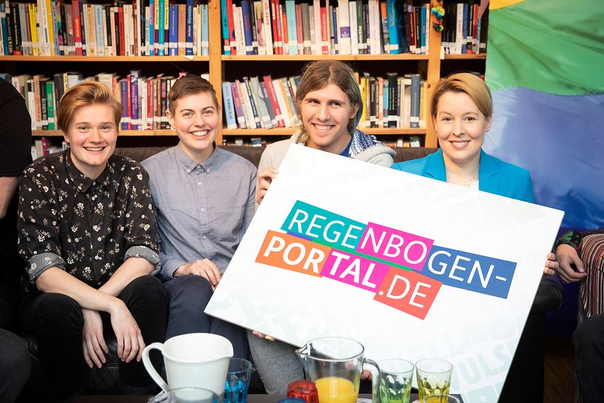 Franziska Giffey mit jungen Leuten vor einer Bücherwand. Sie halten ein Schild hoch mit der Aufschrift regenbogenportal.de