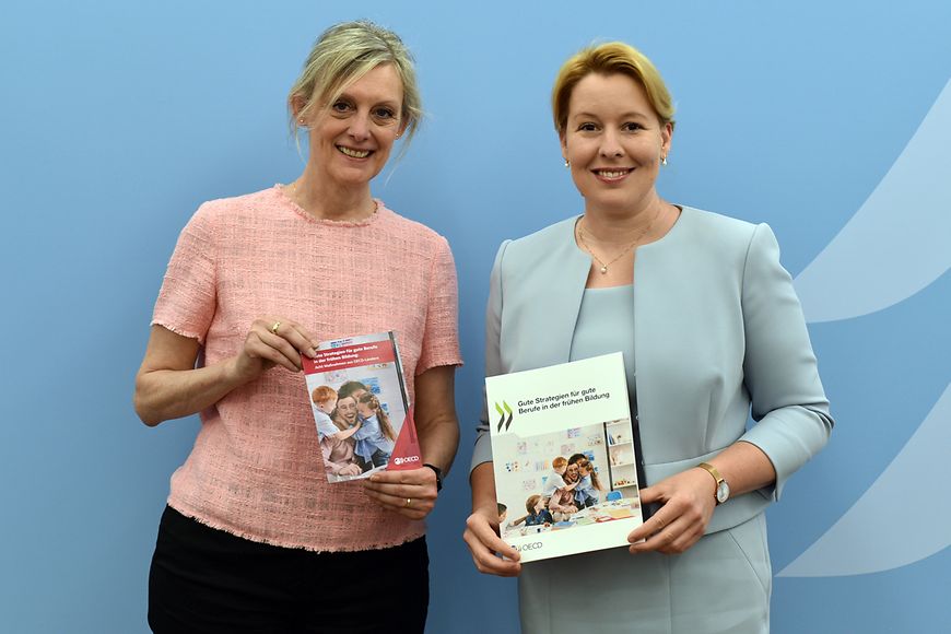 Franziska Giffey und Monika Queisser von der OECD mit der neuen Studie vor einen blauen Wand im Bundesfamilienministerium