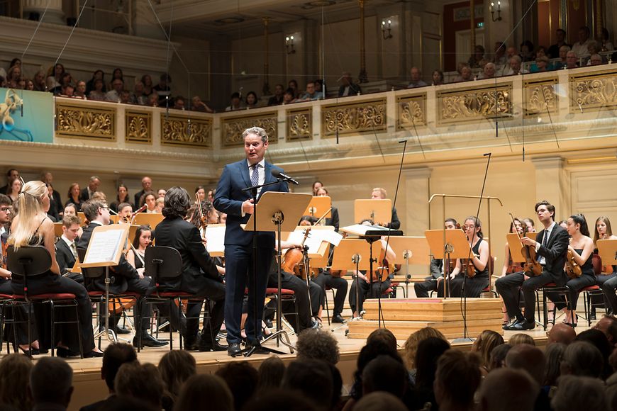 Stefan Zierke am Redepult im vollen Konzertsaal und umringt von zahlreichen Musikerinnen und Musikern