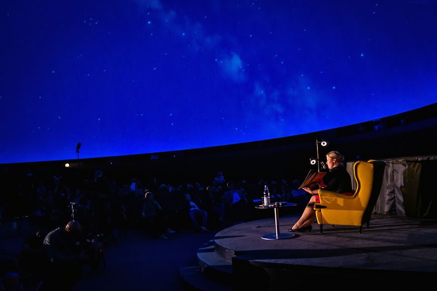 Dr. Franziska Giffey liest auf einer Bühne ein Buch im abgedunkelten Großplanetarium