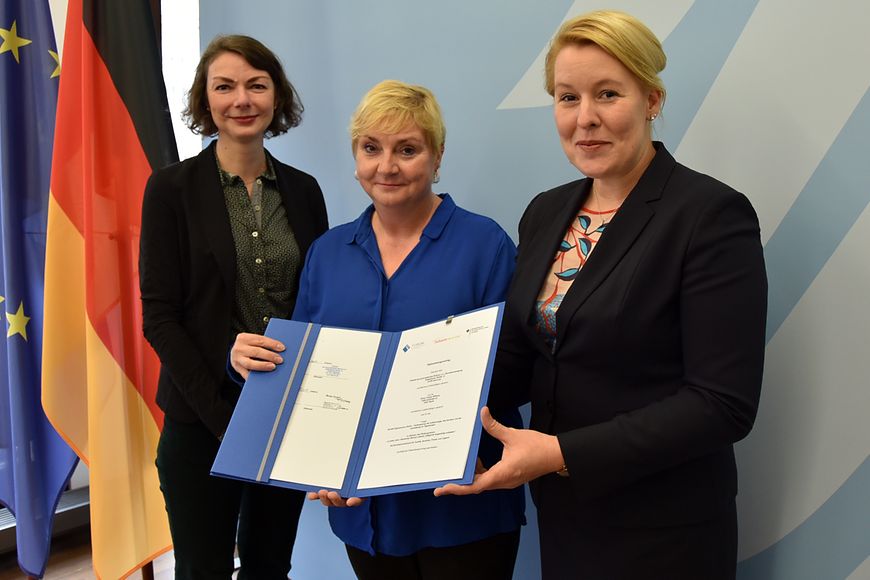 Drei Personen halten eine Förderbewilligung in den Händen vor einer blauen Wand mit der Aufschrift 