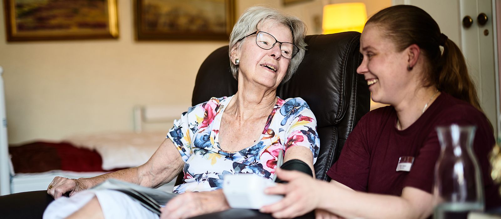 Eine ältere Frau sitzt im Sessel, eine Pflegerin misst den Blutdruck