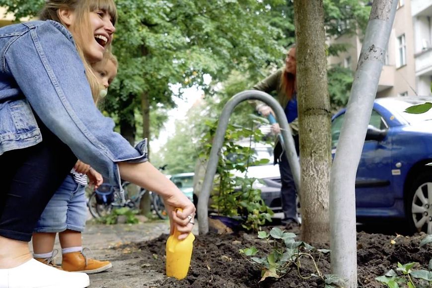 Das Bild zeigt eine Mutter und ein Kind, die an einem Fußweg etwas neben einem Baum pflanzen