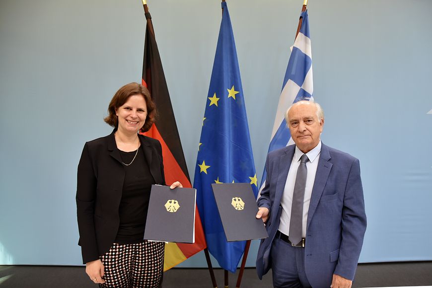 Juliane Seifert und Dr. Georgios Voutsinos vor den Flaggen Deutschland, Griechenland und der Europäischen Union