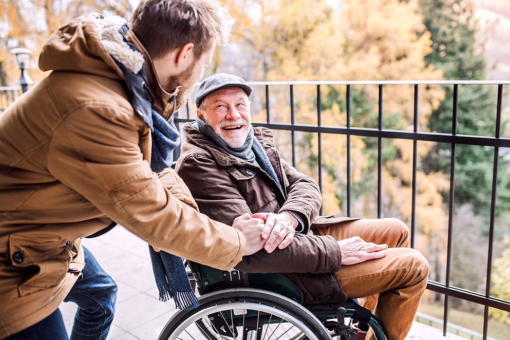 Ein junger Mann besucht einen älteren Mann im Rollstuhl auf dem Balkon