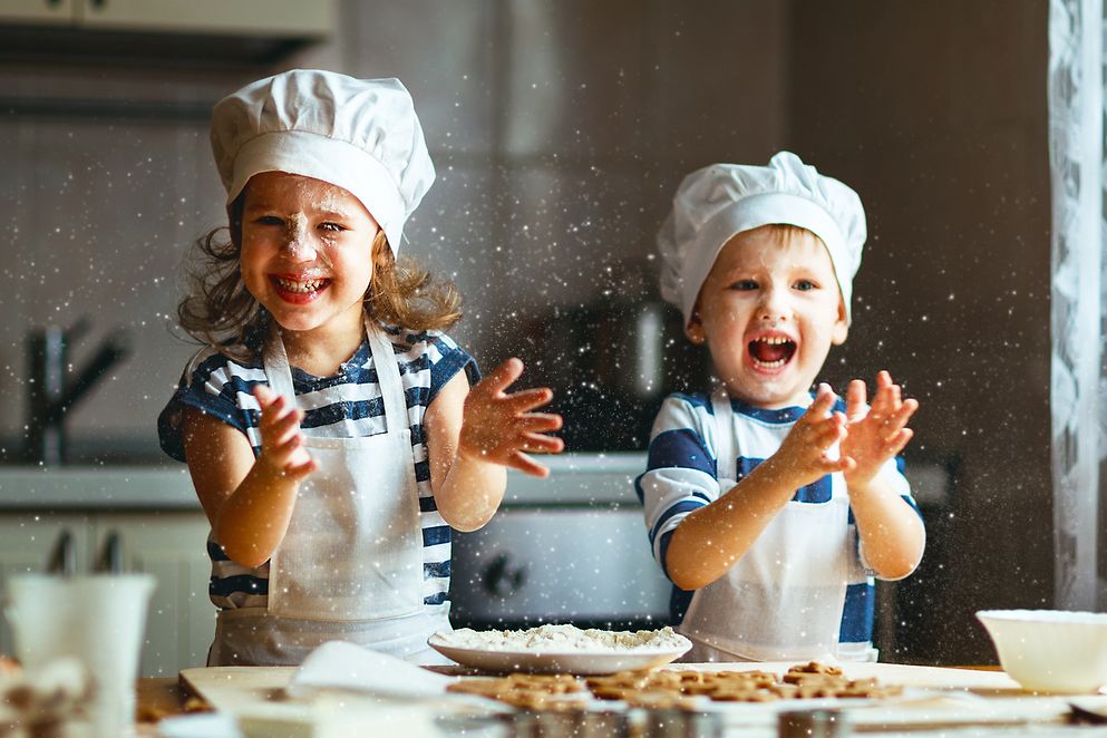 2 Kinder mit Backmützen in einer Küche backen Waffeln und freuen sich