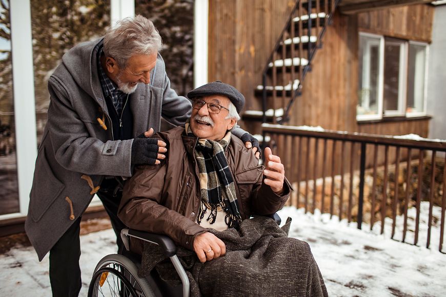 Ein älterer Mann im Rollstuhl wird von seinem Partner geschoben