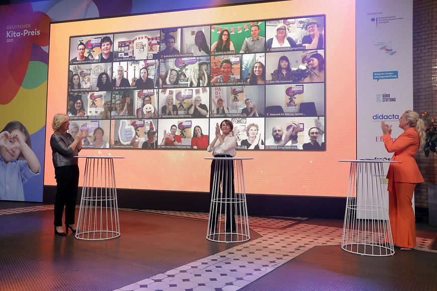 Christine Lambrecht und Elke Büdenbender stehen vor einer Videowand mit den Gewinnerinnen und Gewinnern und klatschen