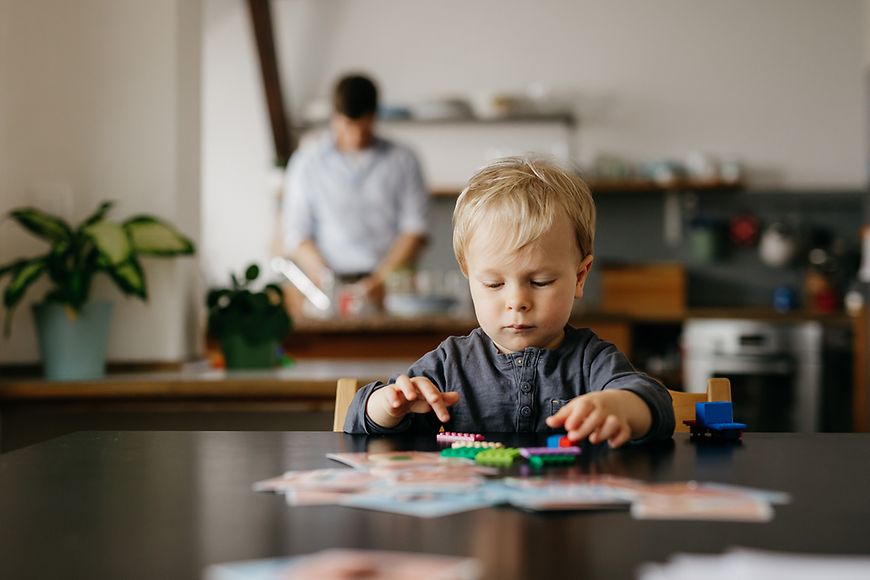 Ein Kind sitzt am Tisch und Spielt, im Hintergrund ist der Vater zu sehen