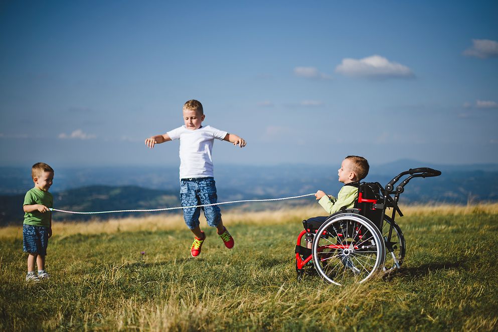 Jungen beim Seilspringen auf einem Feld, einer von ihnen hält im Rollstuhl das Seil
