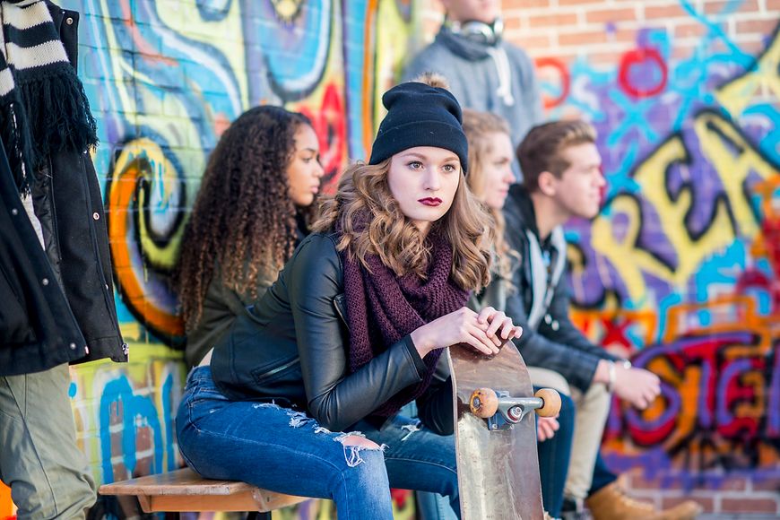 Ein Mädchen mit Skateboard sitzt mit anderen Jugendlichen vor einer bemalten Wand.