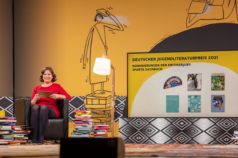 Juliane Seifert gibt neben einem großen Stapel Bücher die Preisträgerinnen und Preisträger bekannt