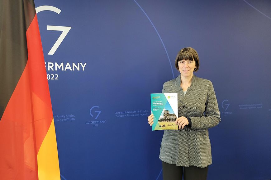 Margit Gottstein mit dem zweiten Bericht des Kompetenzzentrums Jugend-Check vor einer blauen Wand mit D-Flagge