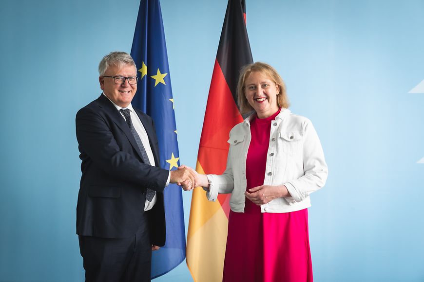Lisa Paus und EU-Kommissar Nicolas Schmit vor einer blauen Wand im Bundesfamilienministerium mit Flaggen