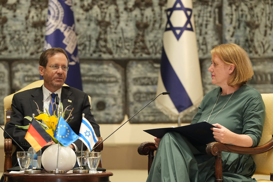 Lisa Paus und Israels Präsident Isaac Herzog sitzen an einem Tisch mit kleinen Flaggen und Blumen und unterhalten sich