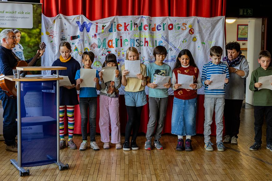 Kinder stehen vor einem Vorgang und singen. Neben ihnen steht ein Mann mit einer Gitarre. 