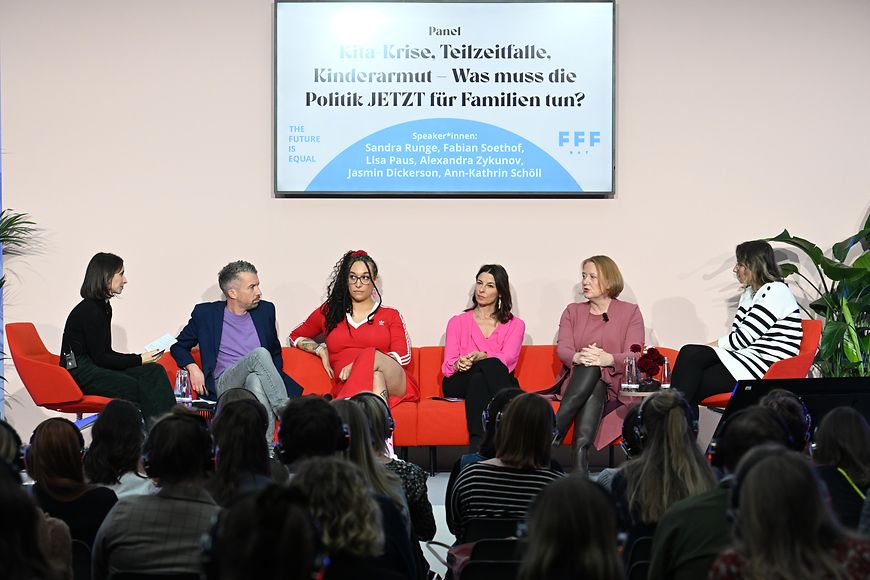 Lisa Paus sitzt mit anderen Diskussionsteilnehmenden auf einem Podium. Vor ihr sitzt das Publikum.