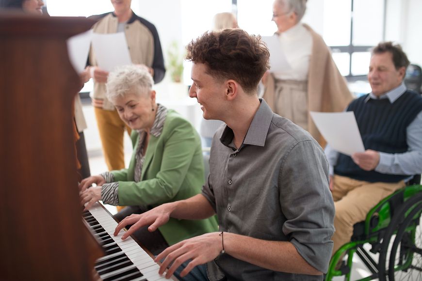 Eine ältere Frau und ein junger Mann spielen gemeinsam Klavier, im Hintergrund singen mehrere Menschen