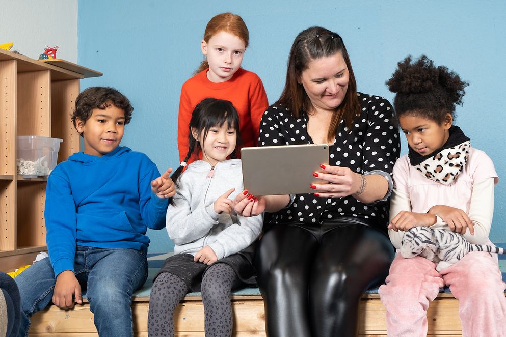 Eine Erzieherin sitzt mit einer Gruppe Kindern auf einer Bank und zeigt etwas auf einem Tablet