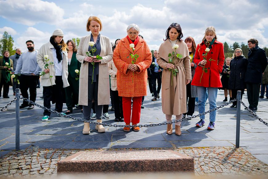 Lisa Paus, Claudia Roth und Annalena Baerbock stehen vor einem Gedenkstein und halten weiße Rosen in der Hand