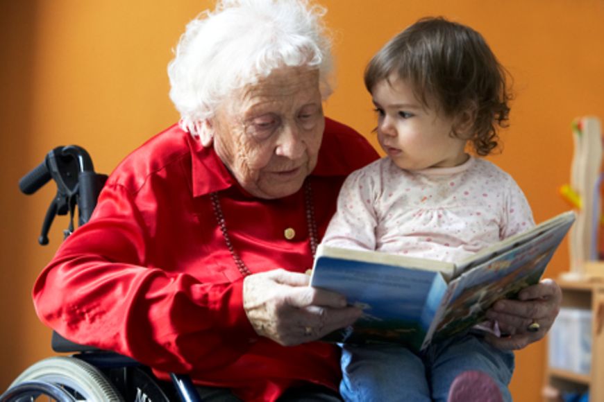 Eine ältere Frau Im Rollstuhl hat ein Kleinkind auf dem Schoß. Sie liest dem Kind aus einem Buch vor
