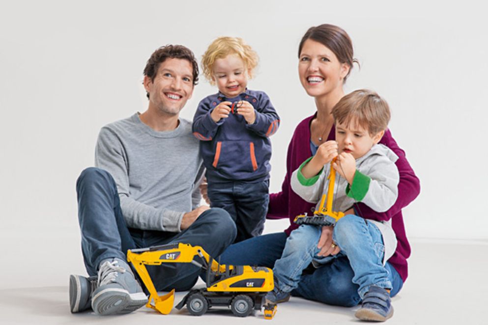 Eine junge Familie sitzt zusammen. Vater, Mutter und die beiden Kinder strahlen und spielen mit Legosteinen.