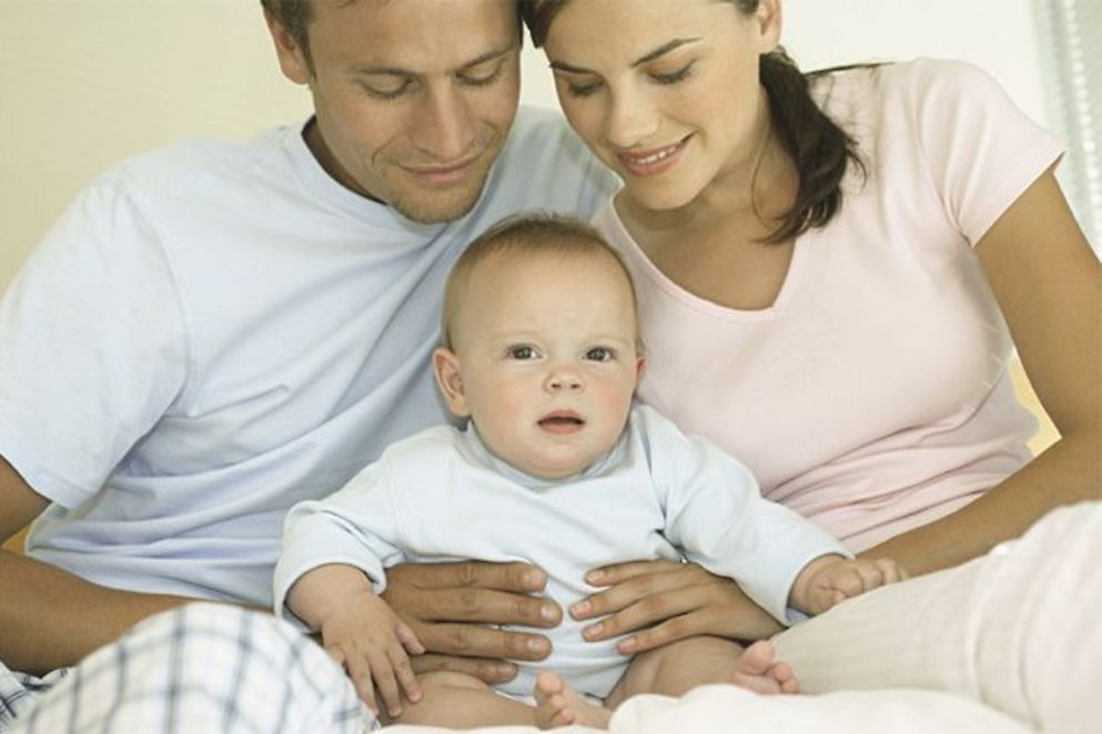 Eine junge Mutter und ein junger Vater sitzen gemeinsam mit ihrem Nachwuchs auf einem Bett.