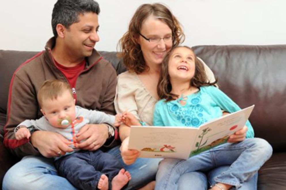 Eine vierköpfige Familie sitzt auf einem Sofa und schaut sich ein Kinderbuch an. 