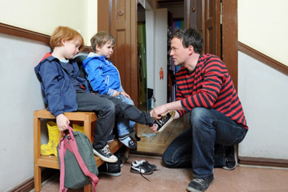 Ein Vater hilft seinen zwei Kindern beim Anziehen Ihrer Schuhe. Bildquelle: M. Gronau / BMFSFJ