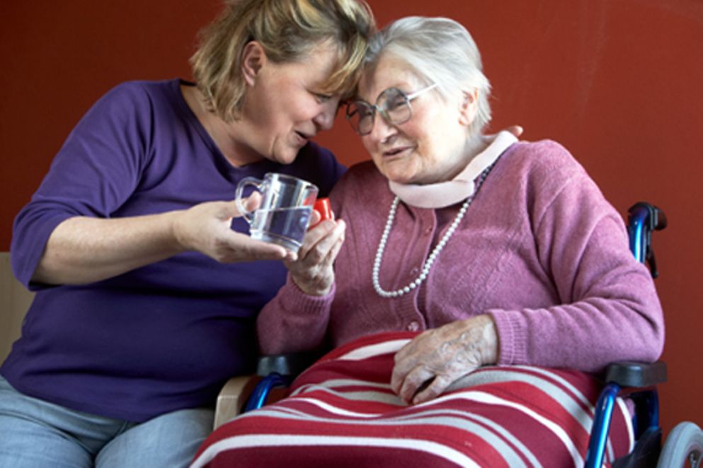 Eine Pflegerin reicht einer älteren Frau im Rollstuhl ein Glas Wasser, Bildnachweis: BMFSFJ / Nagold