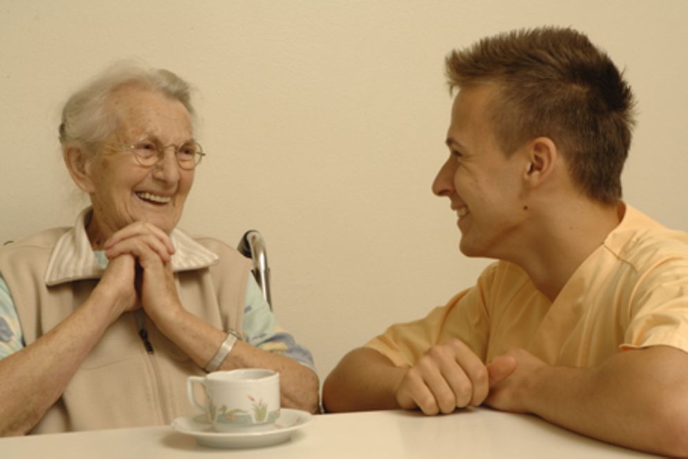 Ein junger Mann und eine alte Frau im Rollstuhl sitzen an einem Tisch. Bildnachweis: BMFSFJ