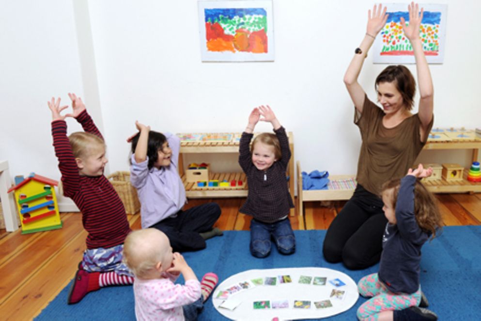 Kinder sitzen zusammen mit einer Erwachsenen im Kreis auf den Boden und heben die Hände. Bildnachweis: Bildnachweis Maike Gronau