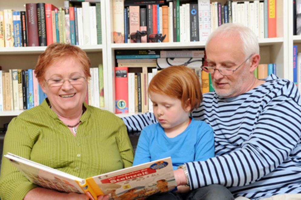 Großeltern sitzen mit ihrer Enkeltochter auf einem Sofa und lesen aus einem Buch. Bildquelle: M. Gronau / BMFSFJ