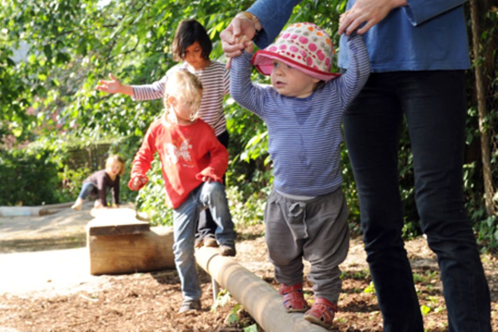 Kinder balancieren mit elterlicher Unterstützung auf einem Holzsteg. 