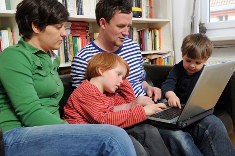 Eine vierköpfige Familie sitzt auf einem Sofa und schaut in einen Laptop